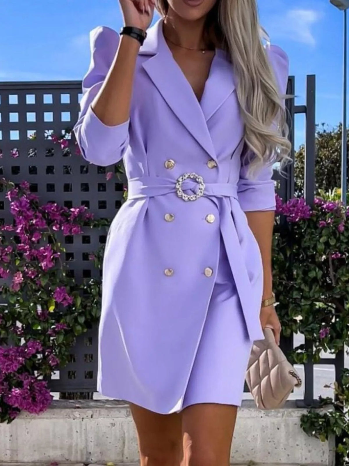 Woman wearing a stylish lilac blazer dress with gold buttons, holding a quilted beige clutch, standing outdoors with flowers, trendy fashion at Shopimist.