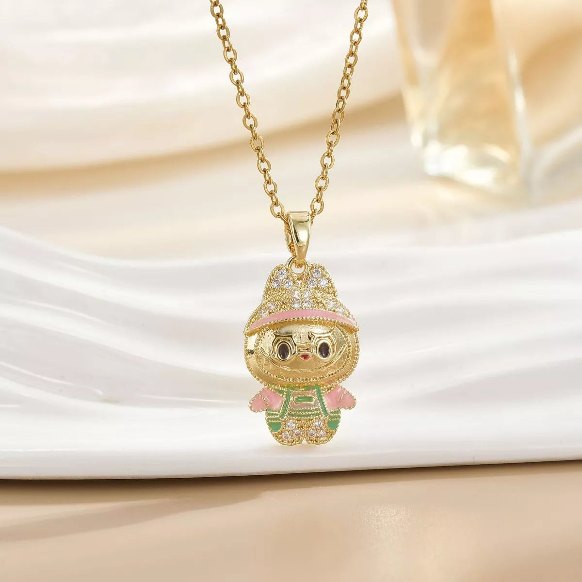 Gold cartoon character pendant necklace with pink and green accents on a light background