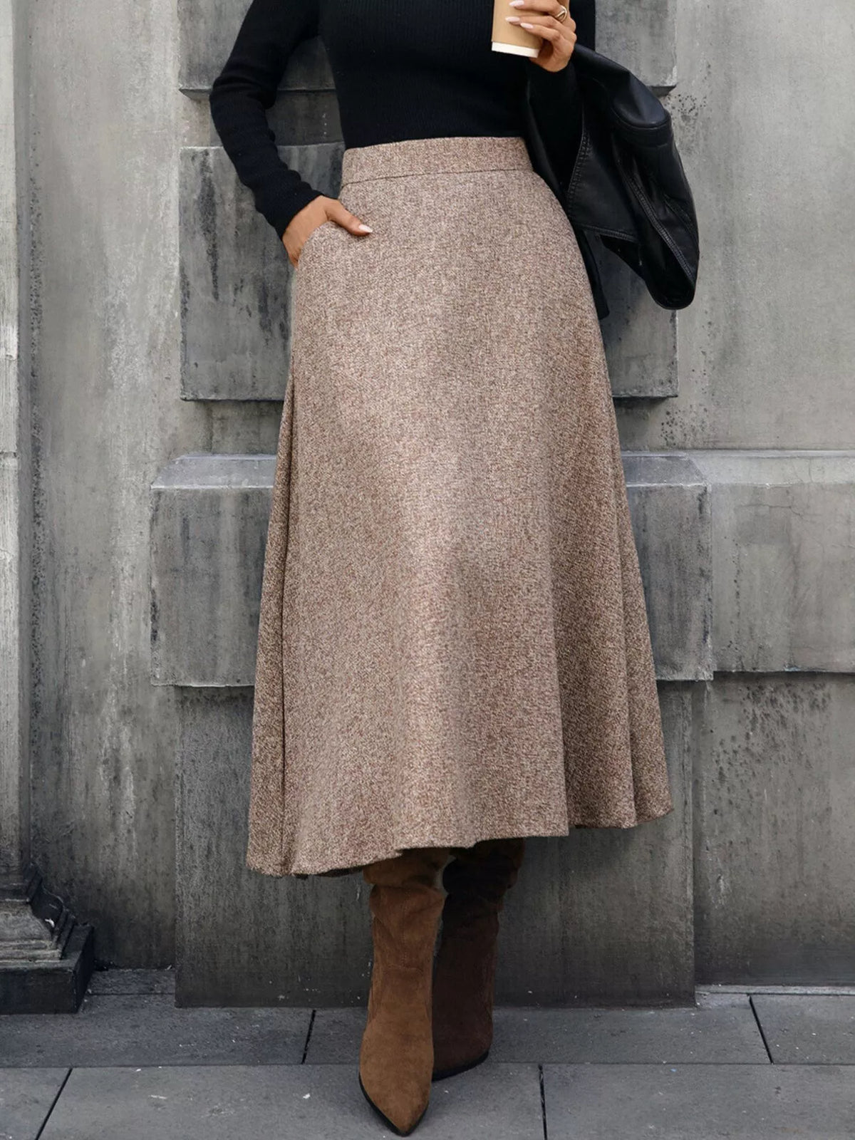 Woman in long brown tweed skirt, black top, and suede boots, holding coffee on city street