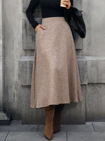 Woman in long brown tweed skirt, black top, and suede boots, holding coffee on city street