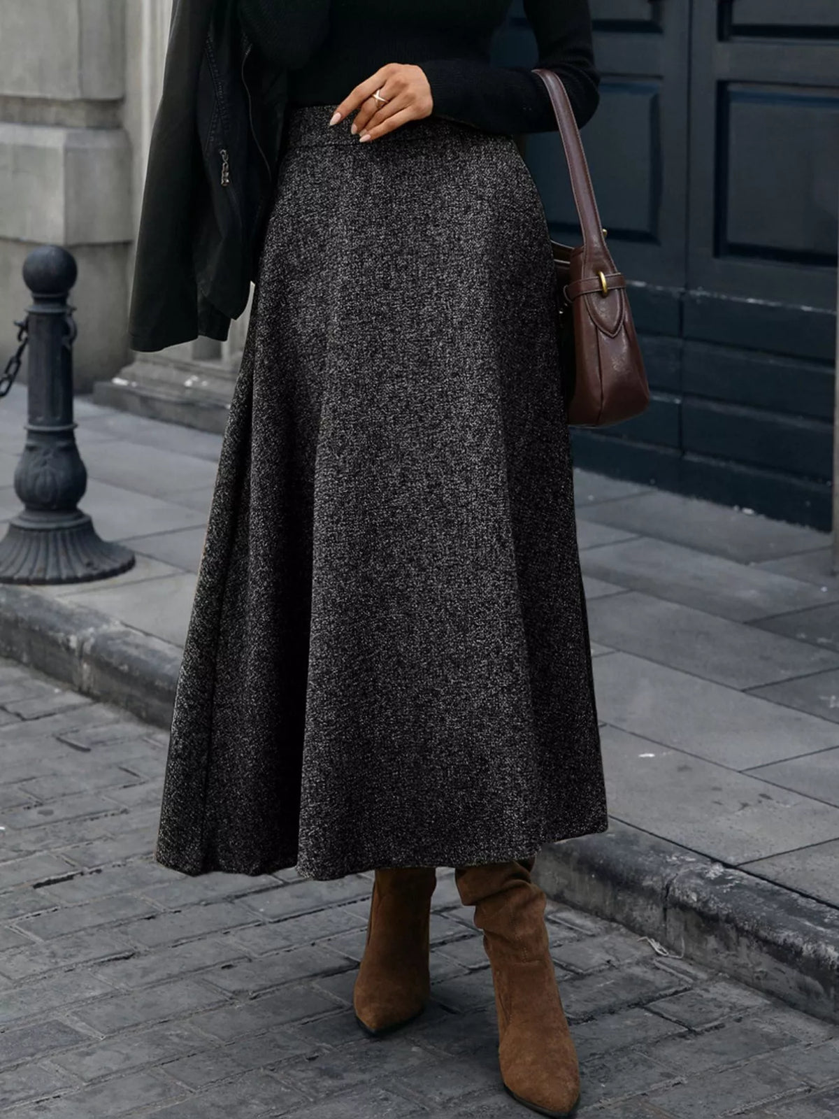 Woman in a black sweater, gray A-line skirt, and brown boots, holding a brown shoulder bag on city street
