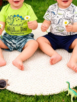 Two toddlers in dinosaur t-shirts sitting on a white mat outdoors, holding daisies, Shopimist kids fashion