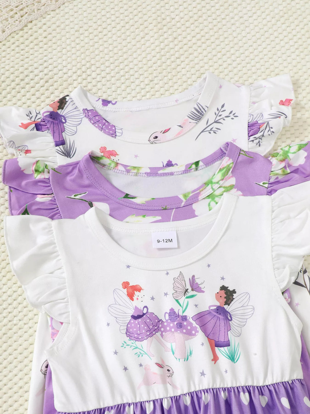 Stack of purple and white baby girl dresses with fairy and floral prints on a textured surface