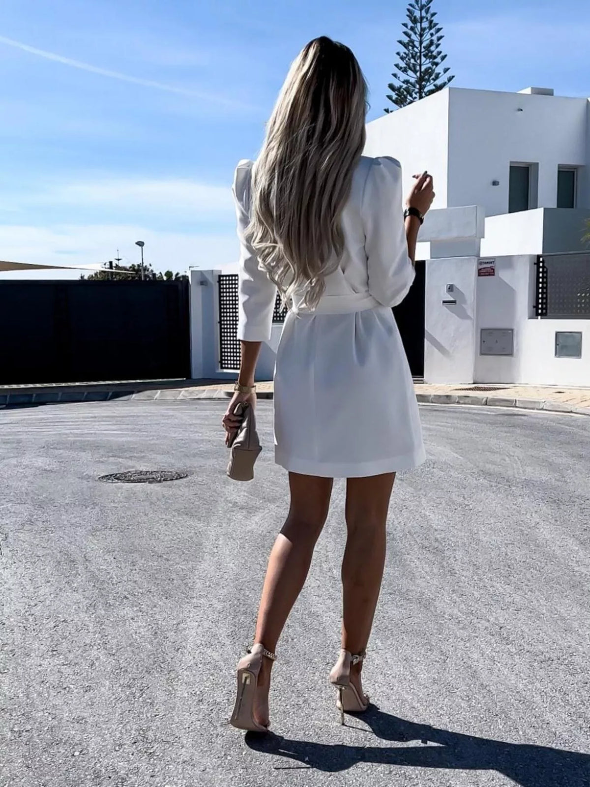 Woman in white dress and heels holding a purse, standing outdoors near a modern white building.