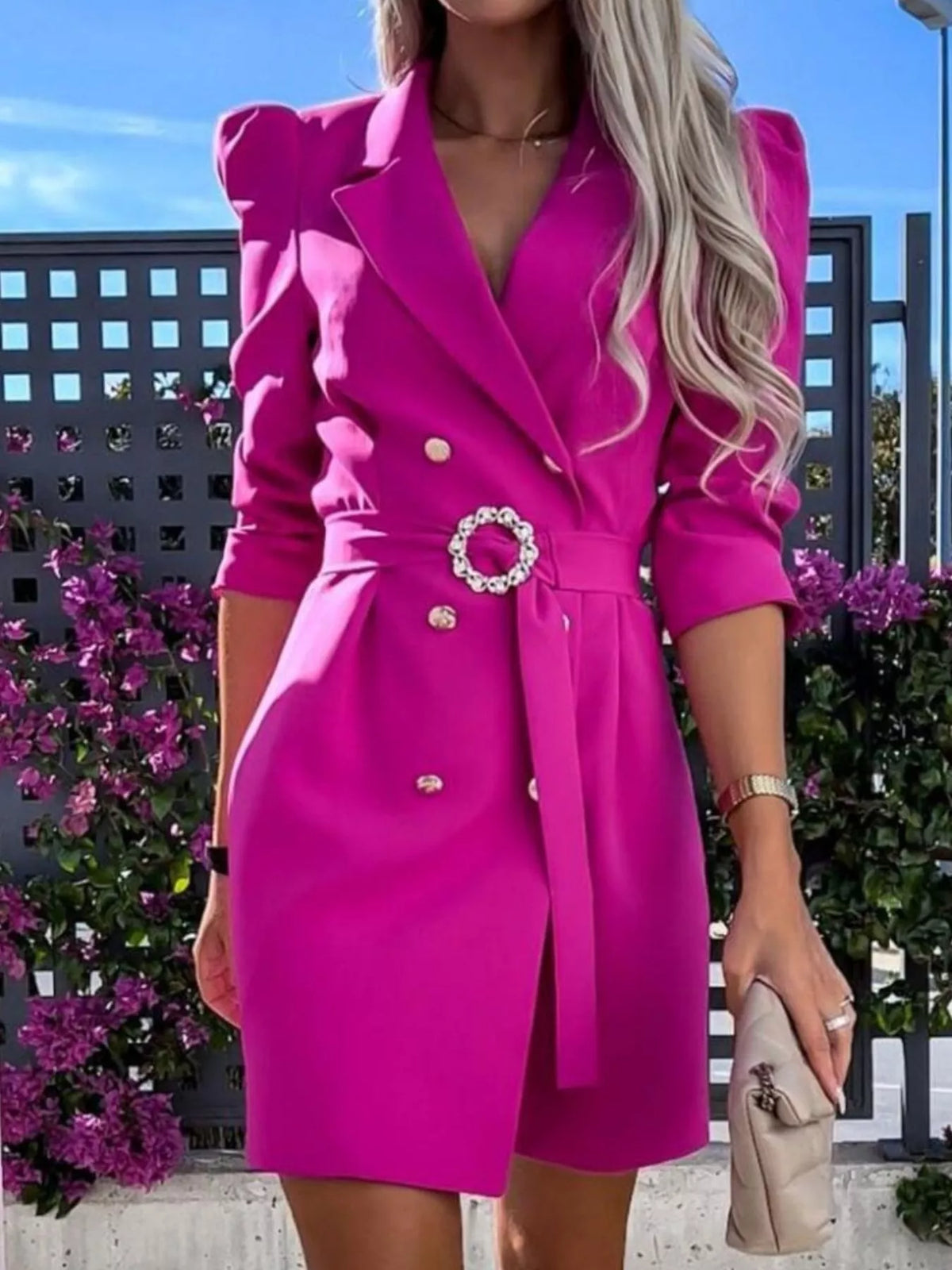 Woman in a bright pink belted blazer dress with gold buttons, holding a beige clutch, outdoor setting