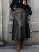Woman wearing black long-sleeve top, gray tweed maxi skirt, brown boots, and holding coffee
