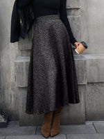 Woman in black sweater and gray maxi skirt with brown boots, holding coffee, stylish street fashion